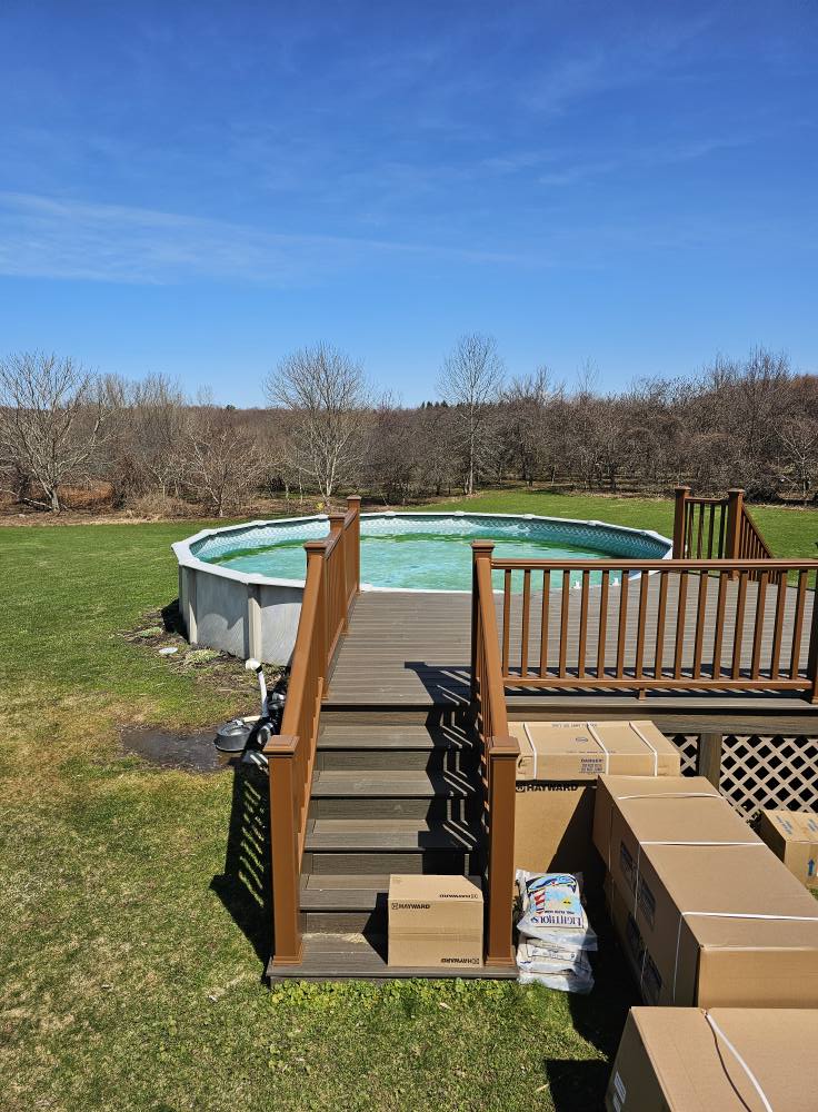 A backyard pool with green water is visible, surrounded by grass and wooden decking. Boxes are stacked near the pool, indicating preparation for maintenance or cleaning.