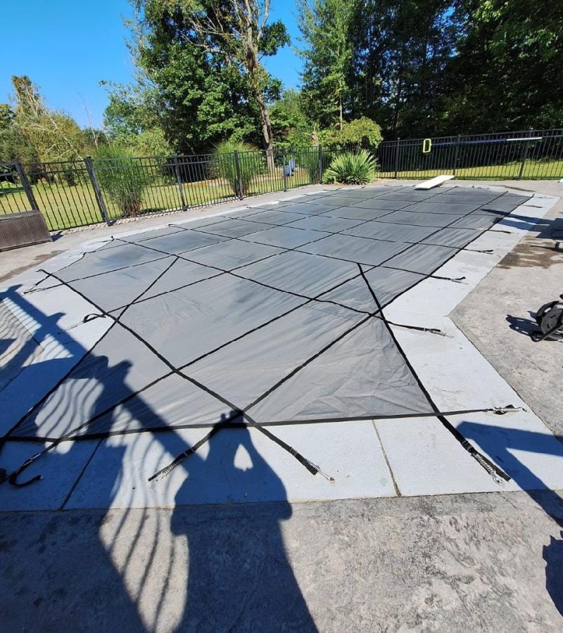 A pool cover is laid out on a concrete deck, with shadows visible from nearby structures. Lush greenery and a fence are seen in the background under clear blue skies.