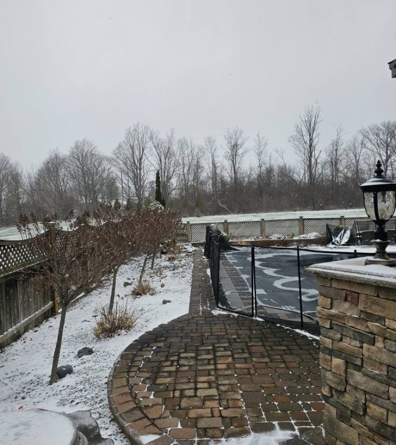 A snowy outdoor scene featuring a curved stone pathway leading to a fenced area, with bare trees in the background and a cloudy sky overhead. Snow covers the ground and some landscaping elements.