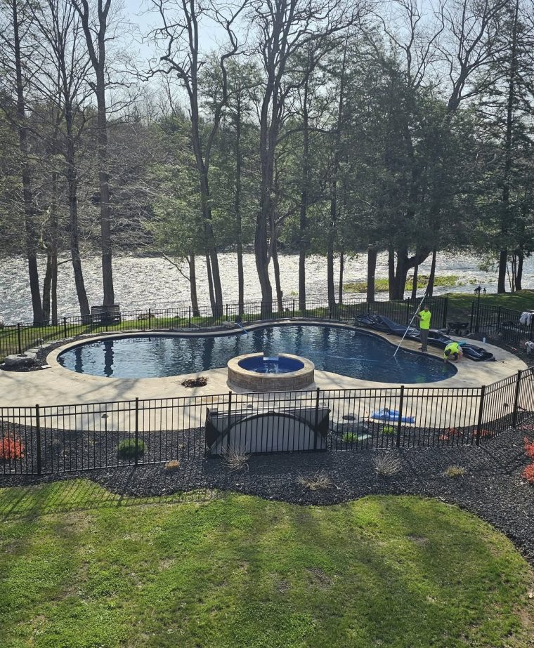 A backyard pool area features a circular pool with a hot tub, surrounded by a black fence and landscaped with flowers and stones. Trees and a river are visible in the background.