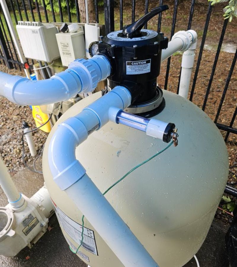 A large round pool filter tank with connected blue pipes and a black motor on top, situated outdoors near a fence. The surrounding area includes some electrical equipment.