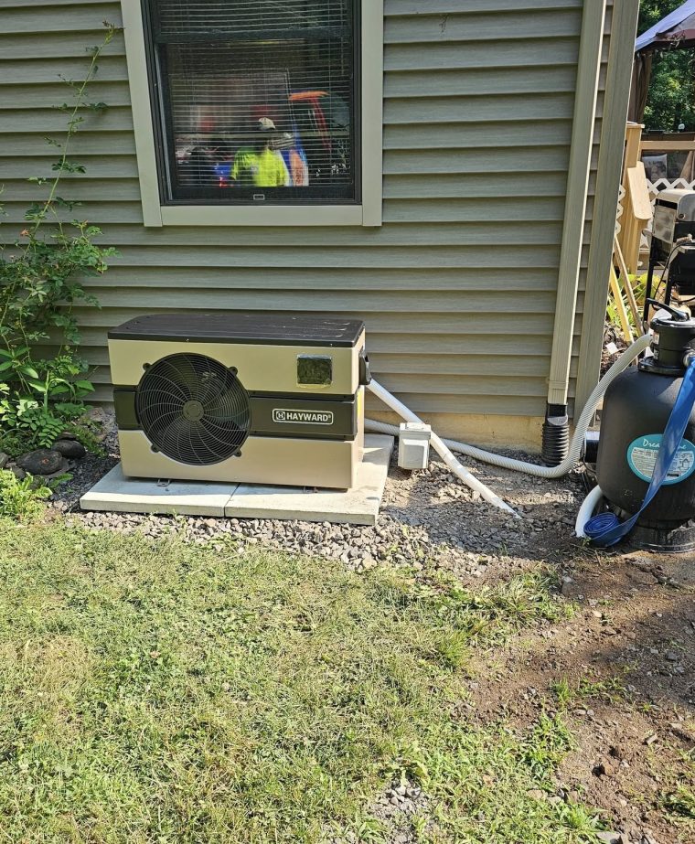 A cooling unit is installed next to a house, with a hose connected to a nearby pump. The area is grassy with some landscaping visible.