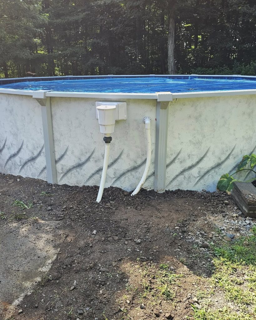 A round above-ground swimming pool is set up on a grassy area, with a ladder on one side and plumbing visible at the bottom. Surrounding the pool are patches of dirt and some plants.