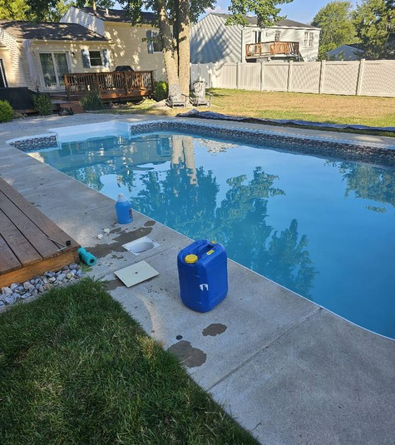 A clear swimming pool is surrounded by a concrete deck and grass, with a blue container and a white object placed nearby. In the background, a house and a fence are visible.