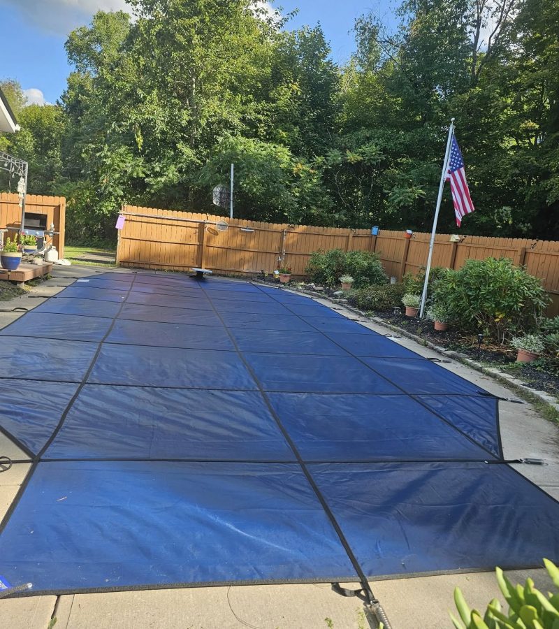 A blue pool cover is spread out over a concrete patio surrounded by greenery and a wooden fence, with an American flag visible in the background.