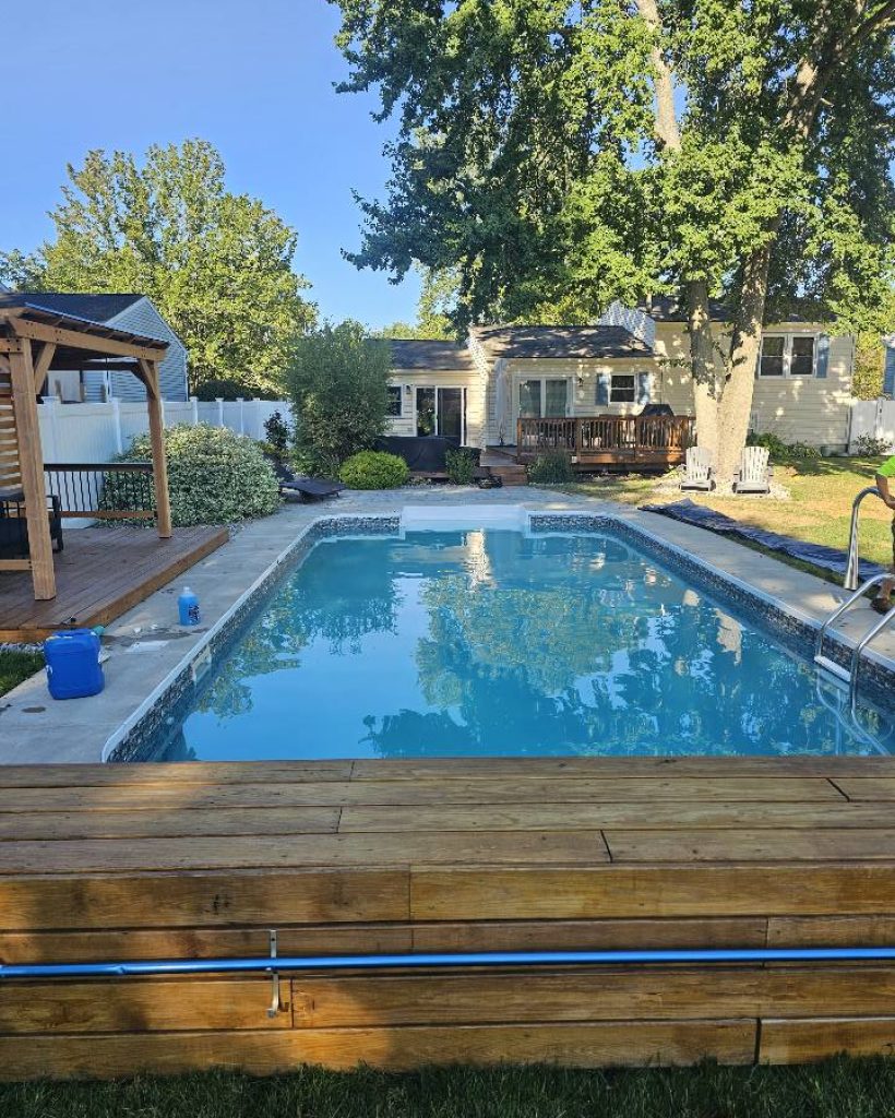 A serene backyard features a clear blue swimming pool surrounded by a wooden deck. A cozy house and lush trees provide a peaceful, inviting atmosphere.