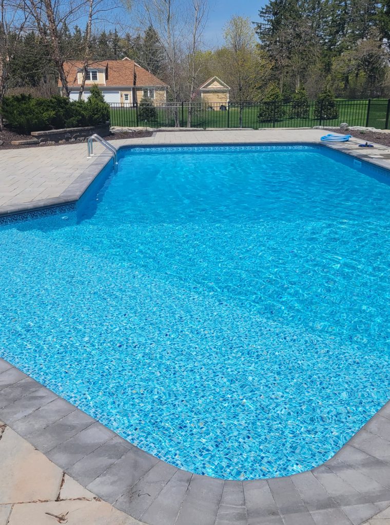 Clear blue swimming pool with stone-paved edges, surrounded by greenery and a residential home. The sunny, serene setting conveys relaxation.